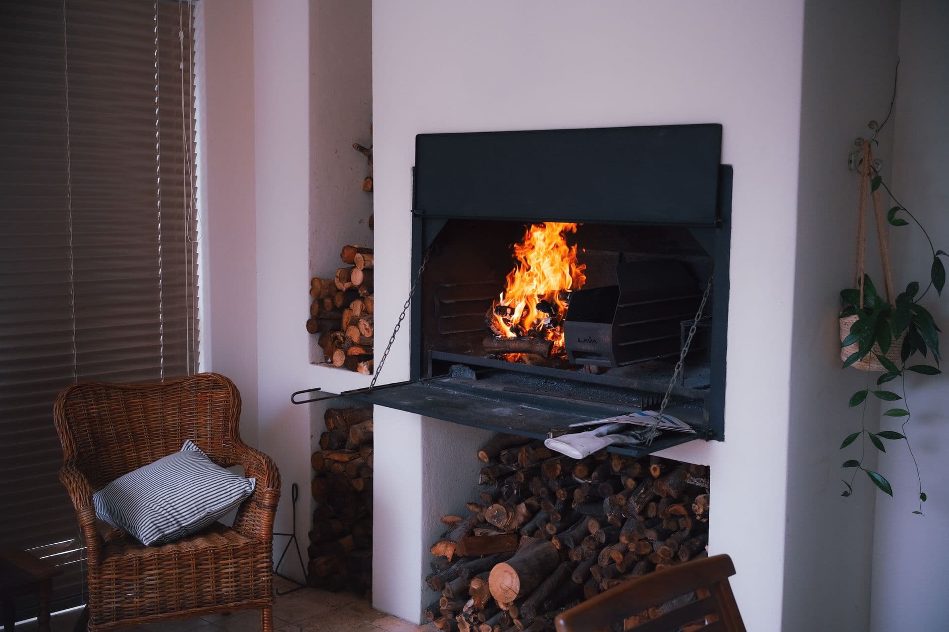 A warm and inviting indoor fireplace with stacked firewood, wicker chair, and plants for a cozy ambiance.